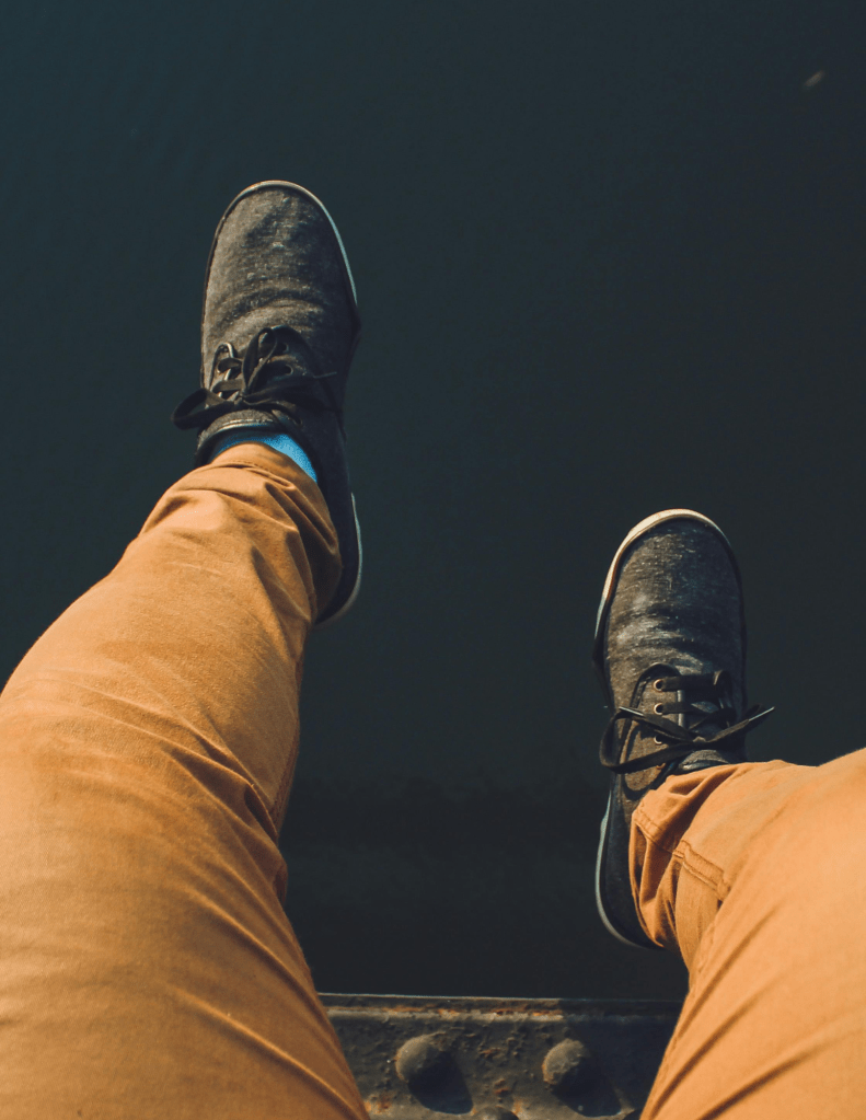 Person wearing brown jeans With a pair of black sneakers sitting on a rail over water