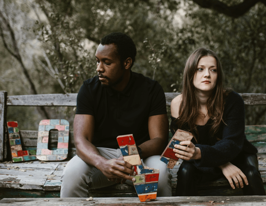 A couple sits in tension on a bench surrounded by nature, holding colorful blocks spelling 'Love'.